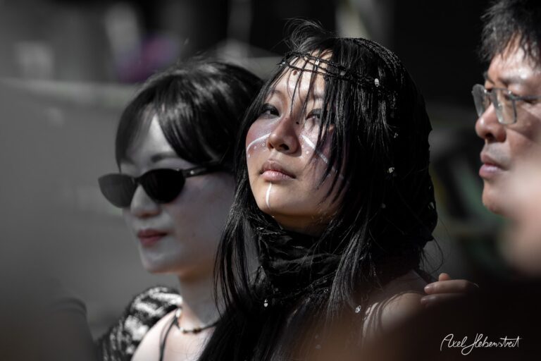Die nachdenkliche Frau mit ethnischen Gesichtsmustern und schwarzem Haar, umgeben von Festivalatmosphäre. – © Axel Hebenstreit