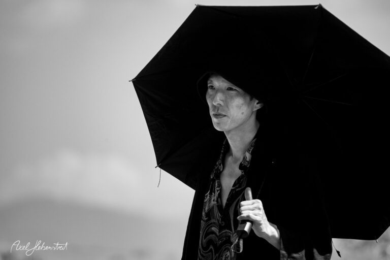 Eine stilvolle Person mit Schirm steht nachdenklich in einer sonnigen Landschaft. – © Axel Hebenstreit