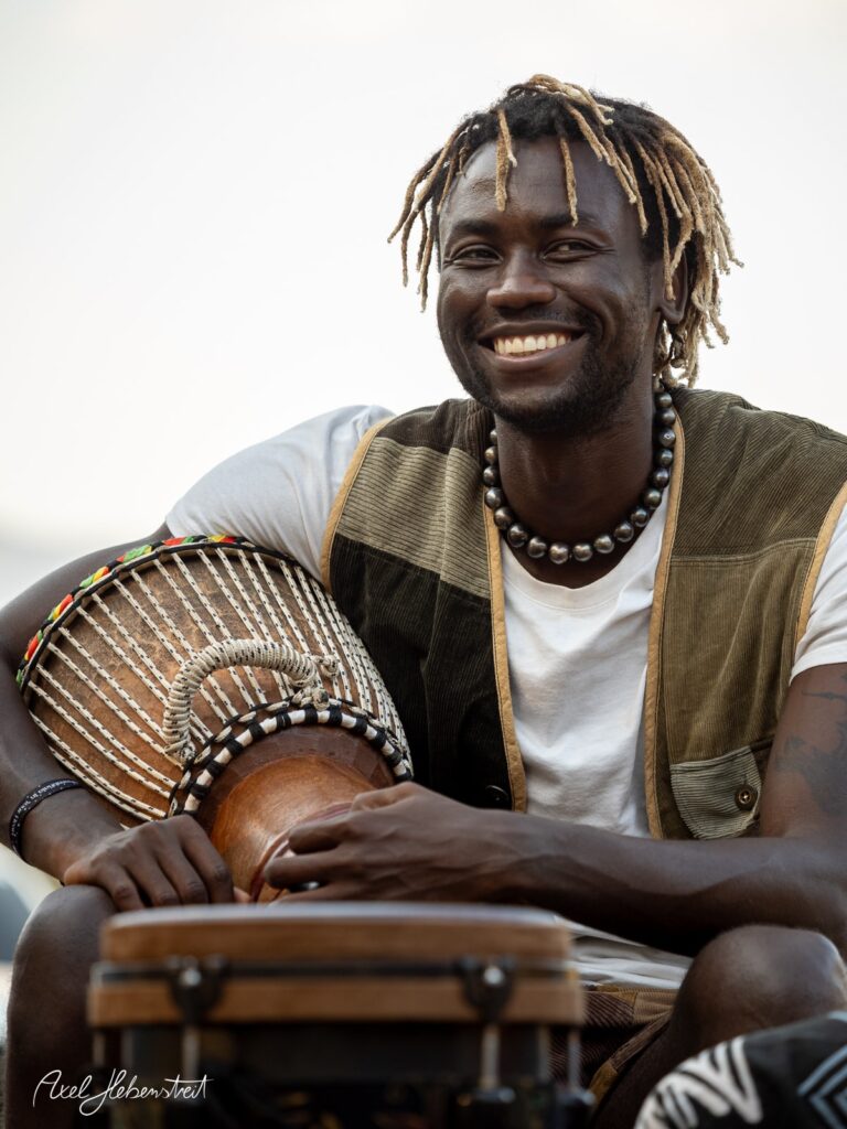 Ein lächelnder Mann mit Dreadlocks spielt auf verschiedenen Trommeln, umgeben von einer entspannten Atmosphäre. – © Axel Hebenstreit