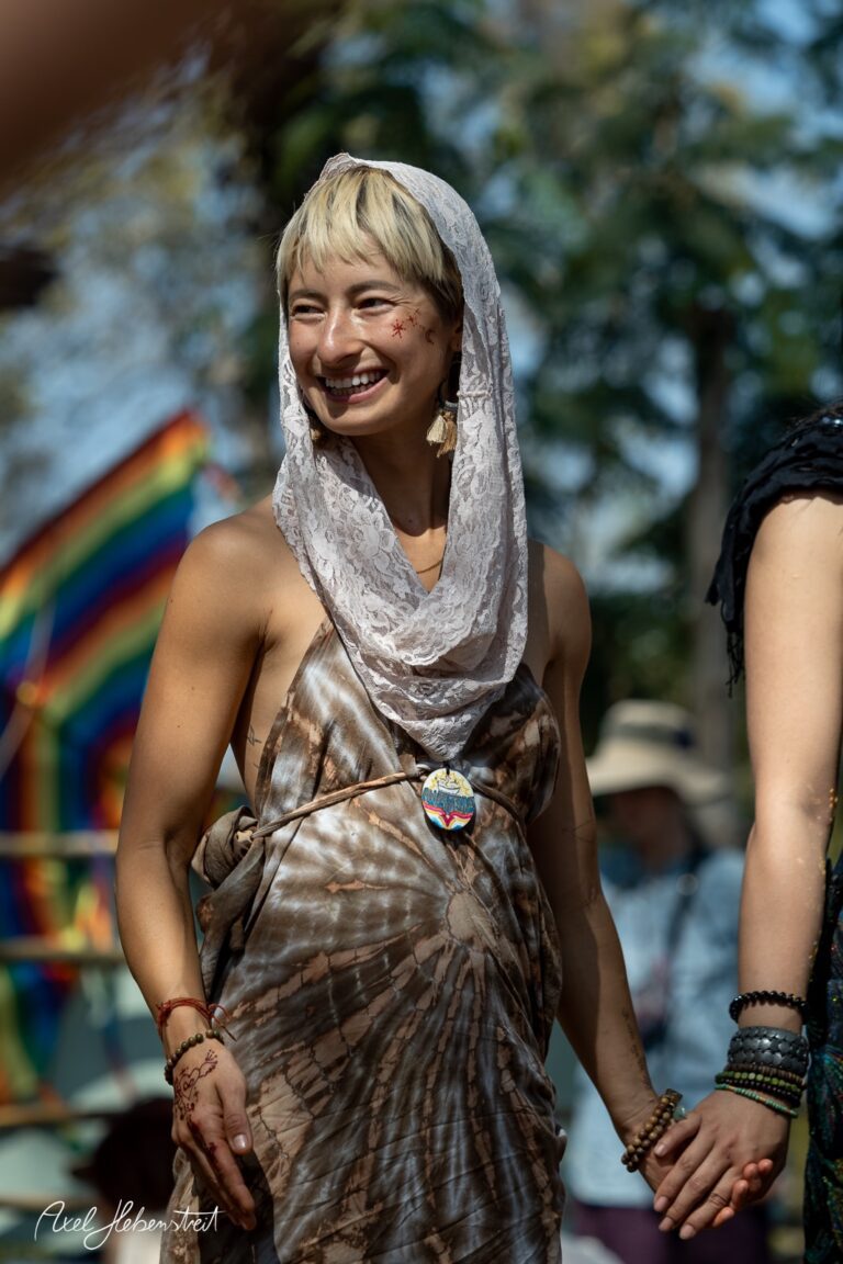 Lächelnde Frau in einem bunten Festival-Outfit, die Händchen haltend mit einer anderen Person eine fröhliche Stimmung vermittelt. – © Axel Hebenstreit
