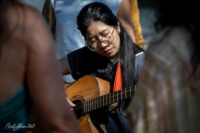 Junge Frau spielt konzentriert Gitarre inmitten einer Gruppe, umgeben von einer entspannten Festivalatmosphäre – © Axel Hebenstreit