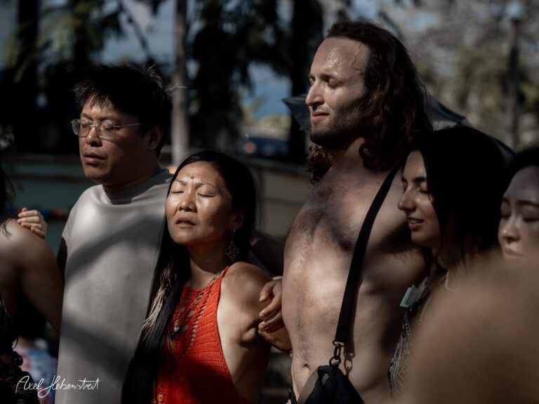 Eine Gruppe von Menschen steht eng beisammen, ihre Augen geschlossen, in einer meditativen Stimmung im Freien – © Axel Hebenstreit