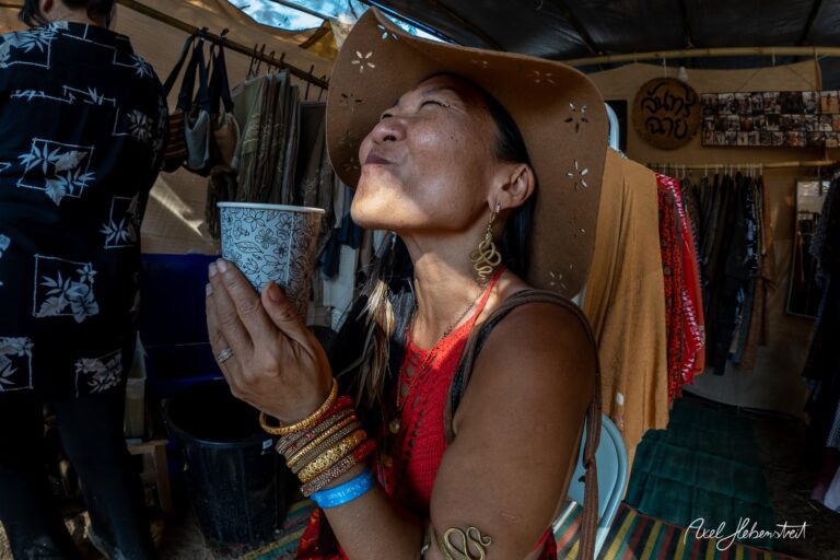 Eine Frau in einem Sonnenhut genießt einen Drink und strahlt Freude aus, umgeben von farbenfrohen Stoffen auf einem Festival. – © Axel Hebenstreit