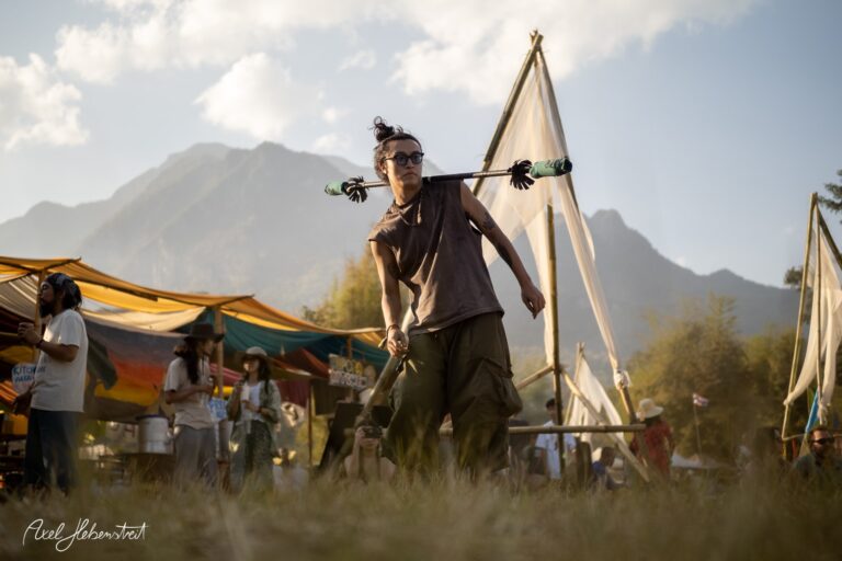 Eine Person mit Pfeilen in der Hand schwingt sie in der grünen Wiese eines Festivals vor Bergkulisse. – © Axel Hebenstreit
