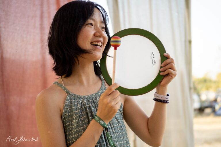 Junge Frau mit einem farbigen Schlaginstrument lächelt glücklich in einem Festival-Ambiente – © Axel Hebenstreit