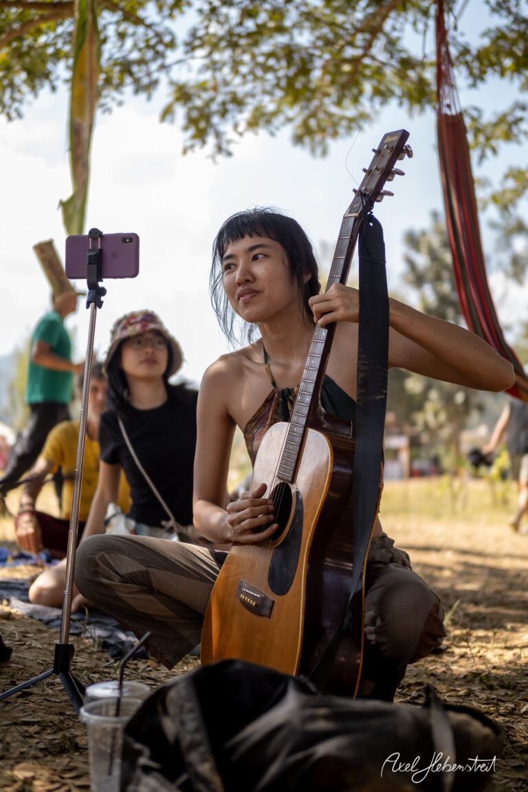 Junge Frau spielt Gitarre und singt unter einem Baum, während sie gefilmt wird – © Axel Hebenstreit