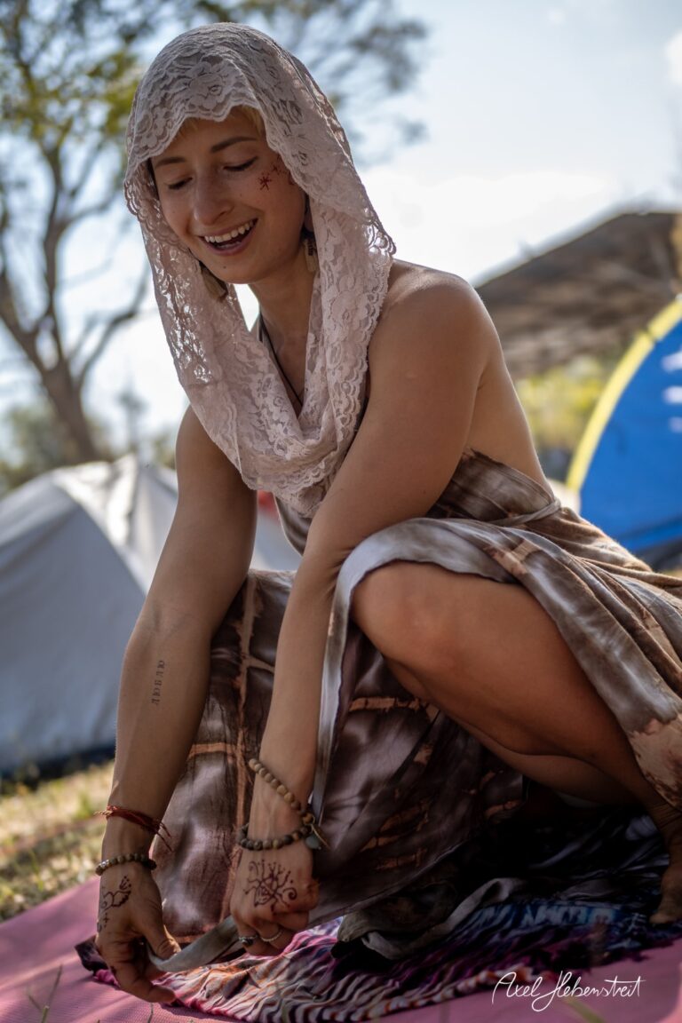 Frau in einem farbenfrohen Kleid sitzt lächelnd auf einer Matte in der Natur und gestaltet mit Leidenschaft. – © Axel Hebenstreit