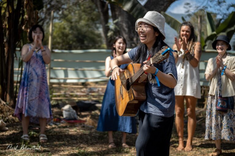 Eine fröhliche Musikerin spielt Gitarre und wird von einer Gruppe applaudierender Menschen umgeben – © Axel Hebenstreit