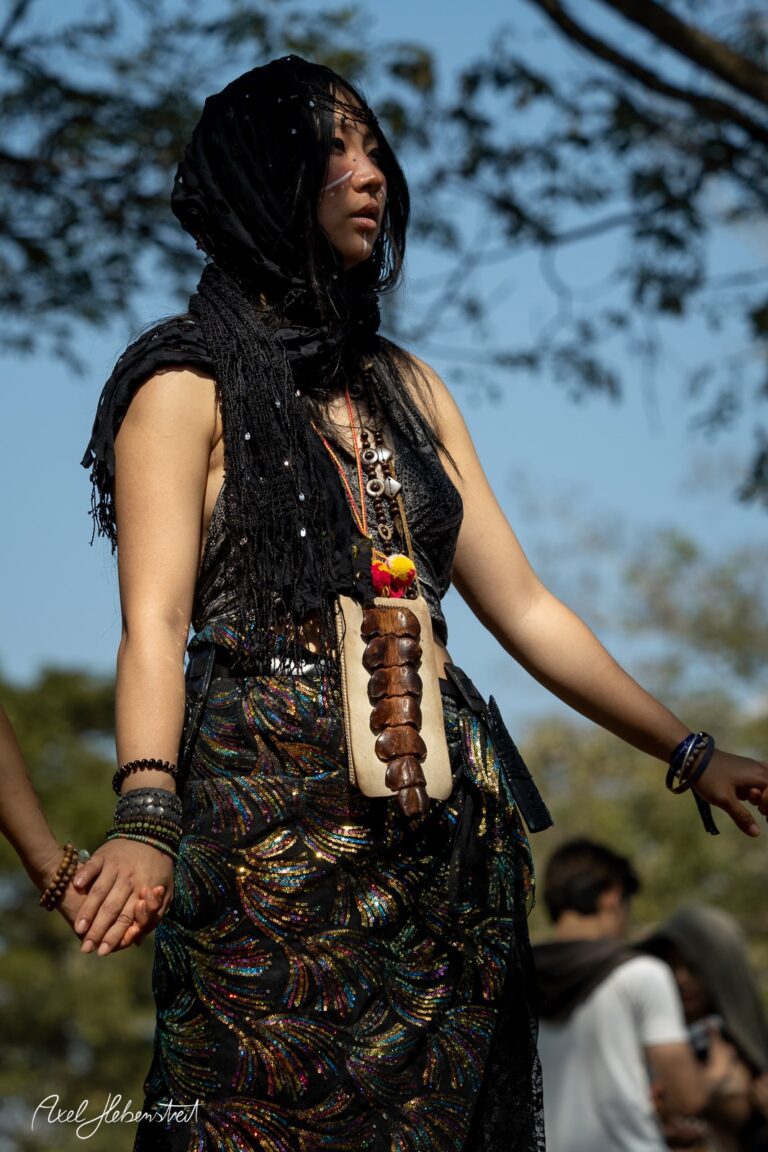 Eine Frau in traditioneller Kleidung mit schwarzem Schal und bunten Halsketten, die in einem sonnigen Park meditiert. – © Axel Hebenstreit