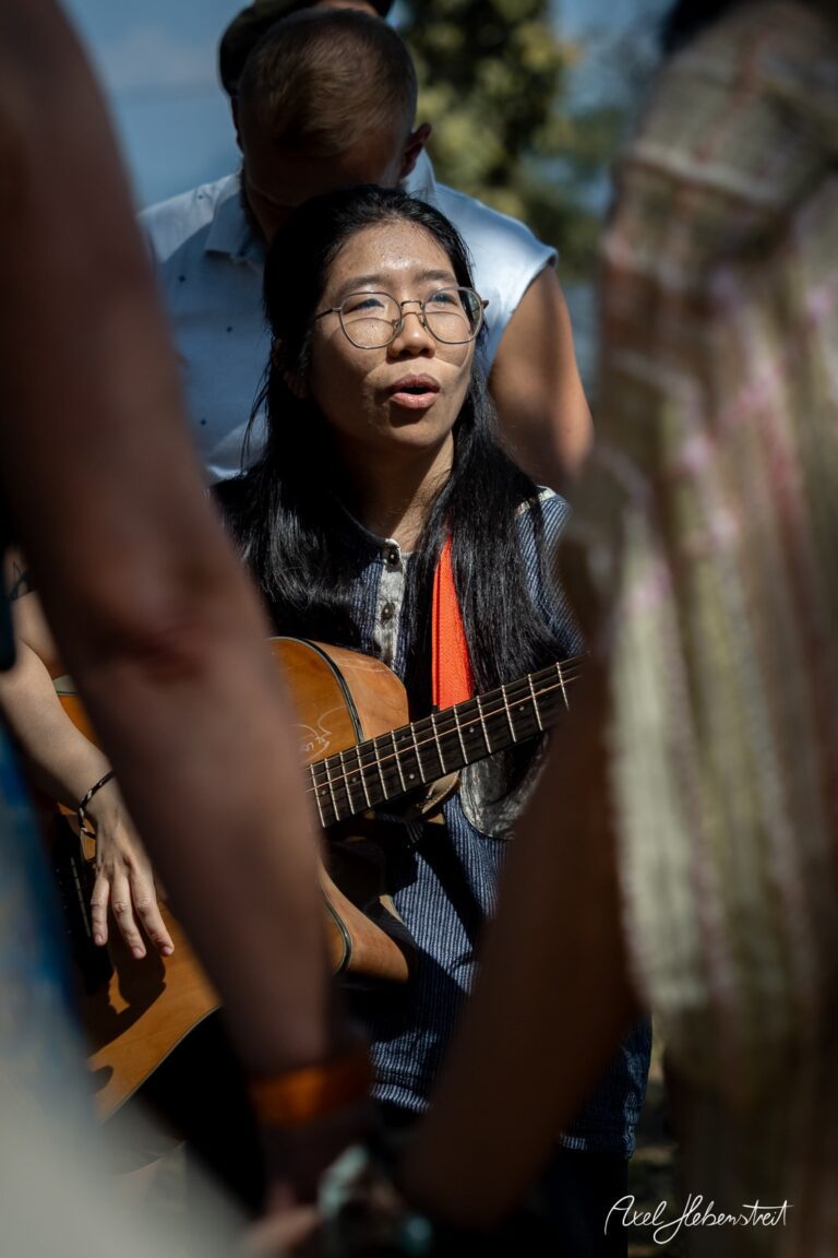 Eine Frau mit Brille spielt Gitarre und singt im Freien, umgeben von Zuhörern in entspannten Kleidern. – © Axel Hebenstreit