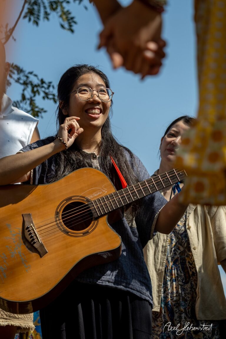 Lächelnde Frau mit Gitarre inmitten einer Gruppe, strahlt Freude und Verbundenheit aus – © Axel Hebenstreit