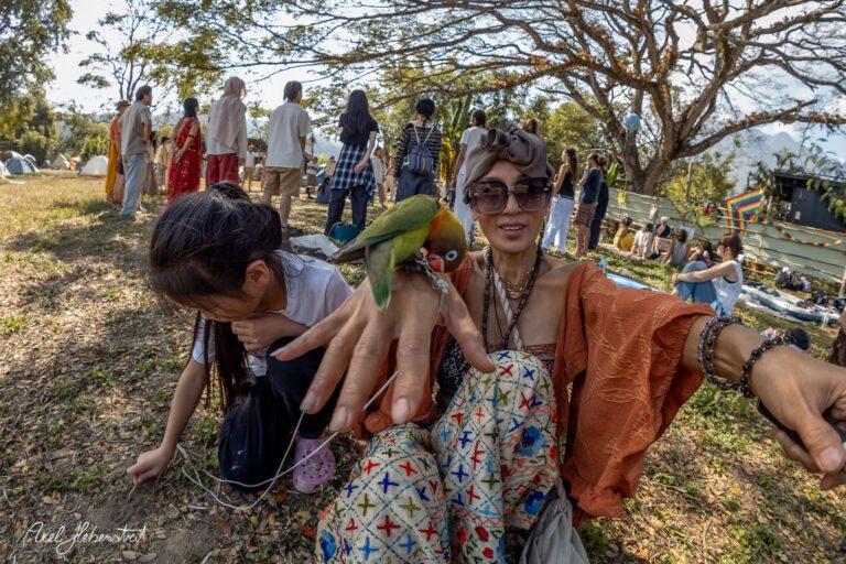 Frau mit bunten Kleidern und einem Papagei auf der Hand, während sie mit einem Kind auf einem Festival sitzt – © Axel Hebenstreit