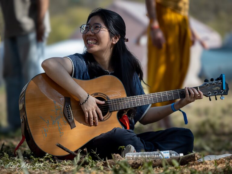 Lächelnde Frau mit Gitarre sitzt auf dem Boden und genießt die Musik im Freien – © Axel Hebenstreit