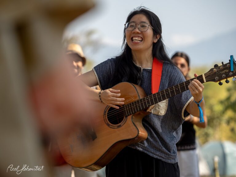 Eine lächelnde Frau spielt Gitarre und singt, umgeben von einer fröhlichen Festivalatmosphäre – © Axel Hebenstreit