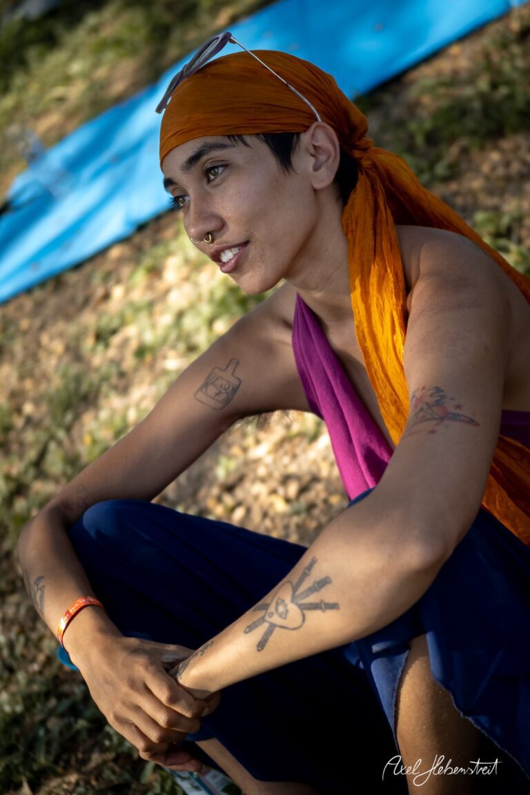 Junge Person mit buntem Kopftuch und Tattoos sitzt nachdenklich im Freien, umgeben von Natur – © Axel Hebenstreit