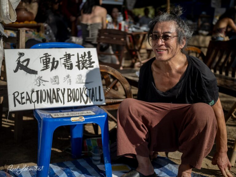 Lächelnder Mann sitzt vor einem Buchstand mit dem Schriftzug „Reactionary Bookstall“ in einer lebhaften Atmosphäre. – © Axel Hebenstreit
