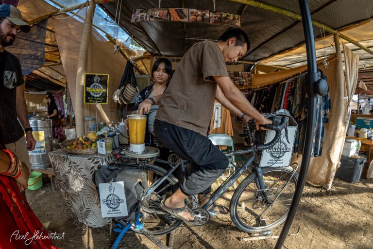 Ein junger Mann bereitet an einem Fahrrad einen Smoothie in einem lebhaften Festivalmarkt – © Axel Hebenstreit