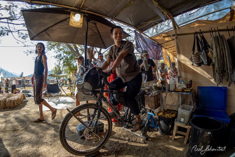 Ein junger Mann fährt auf einem Fahrrad zwischen Ständen auf einem lebhaften Festivalgelände. – © Axel Hebenstreit