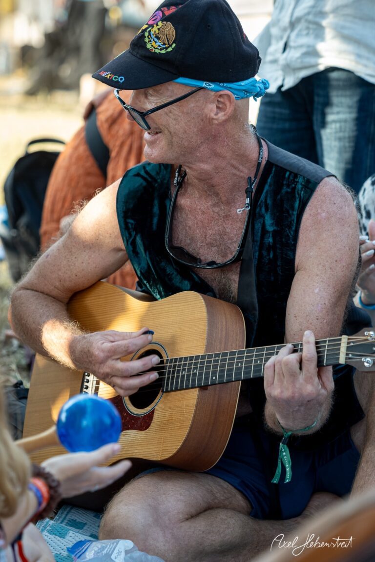 Mann mit einem Gitarrenspiel im Freien, umgeben von einer fröhlichen Festivalatmosphäre – © Axel Hebenstreit