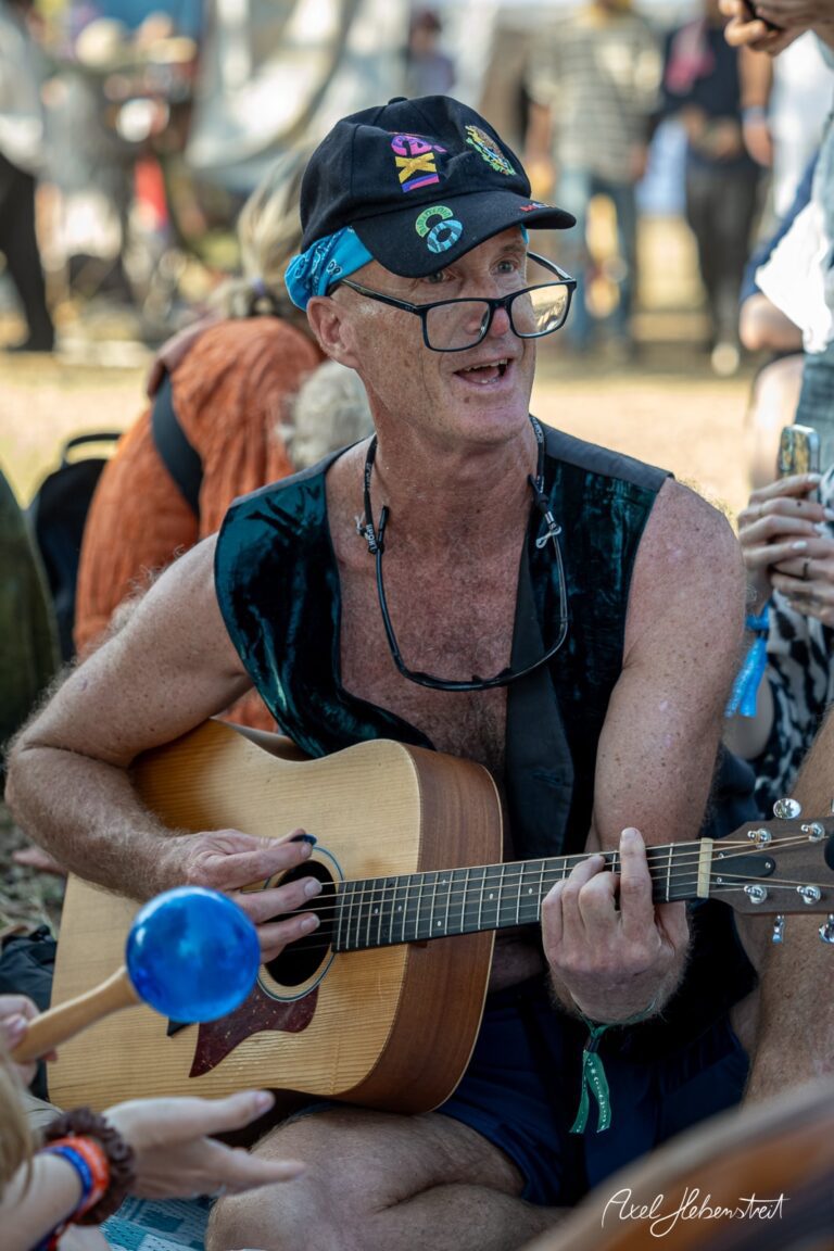Mann mit Brille und Hut spielt Gitarre, umgeben von Menschen beim Festival in ausgelassener Stimmung – © Axel Hebenstreit