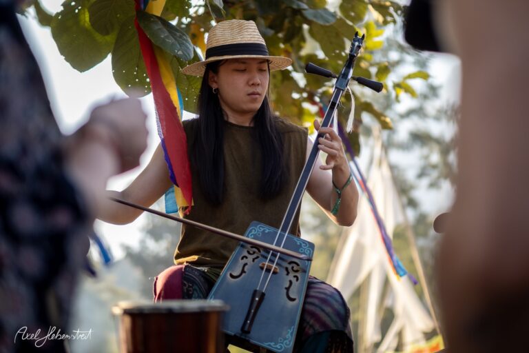 Ein Musiker mit Strohhut spielt ein traditionelles Instrument unter grünen Bäumen während eines Festivals – © Axel Hebenstreit