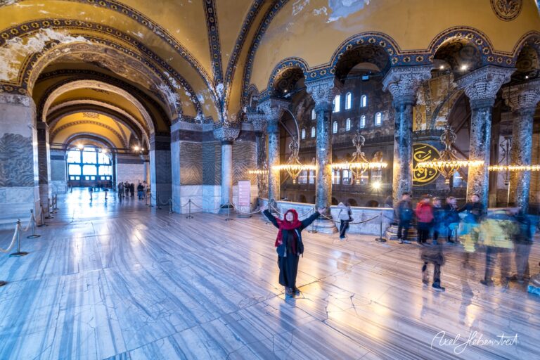 Eine Frau mit rotem Kopftuch breitet die Arme in der beeindruckenden Hagia Sophia aus – © Axel Hebenstreit.