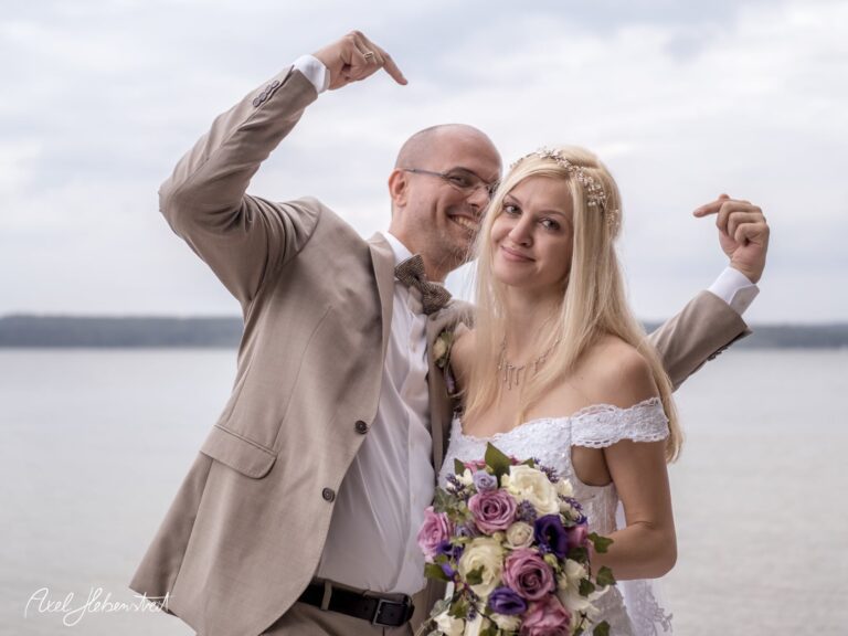 Brautpaar posiert fröhlich am Wasser, umgeben von Hochzeitsblumen, in festlicher Stimmung. – © Axel Hebenstreit