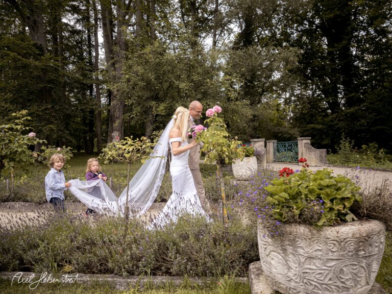 Brautpaar im Hochzeitskleid und Anzug, begleitet von Kindern in einem blühenden Garten – © Axel Hebenstreit