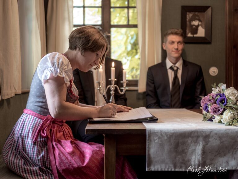 Eine Frau in traditioneller Tracht sitzt an einem Tisch und unterschreibt, während ein Mann im Anzug zuschaut. – © Axel Hebenstreit