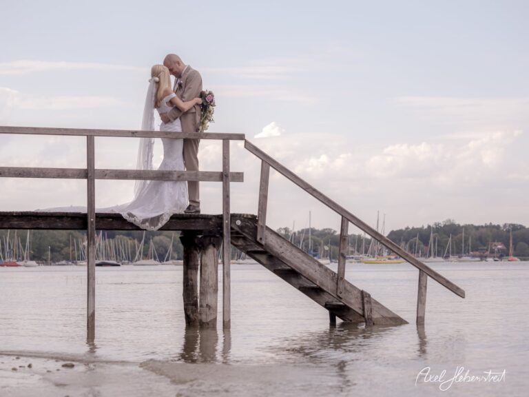 Ein Hochzeitspaar umarmt sich auf einem Holzsteg über dem Wasser, umgeben von einer romantischen Atmosphäre – © Axel Hebenstreit