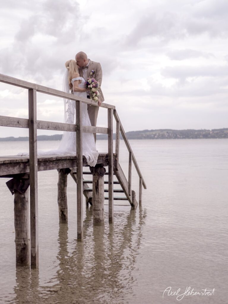 Braut und Bräutigam küssen sich auf einem Steg am Wasser, umgeben von einer romantischen Stimmung – © Axel Hebenstreit