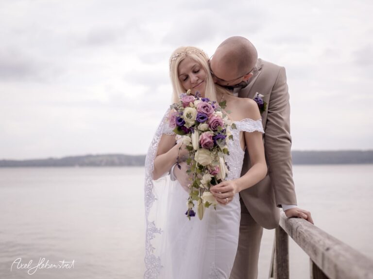 Braut und Bräutigam umarmt am Ufer, umgeben von romantischen Blumen, strahlen Liebe und Zärtlichkeit aus – © Axel Hebenstreit.