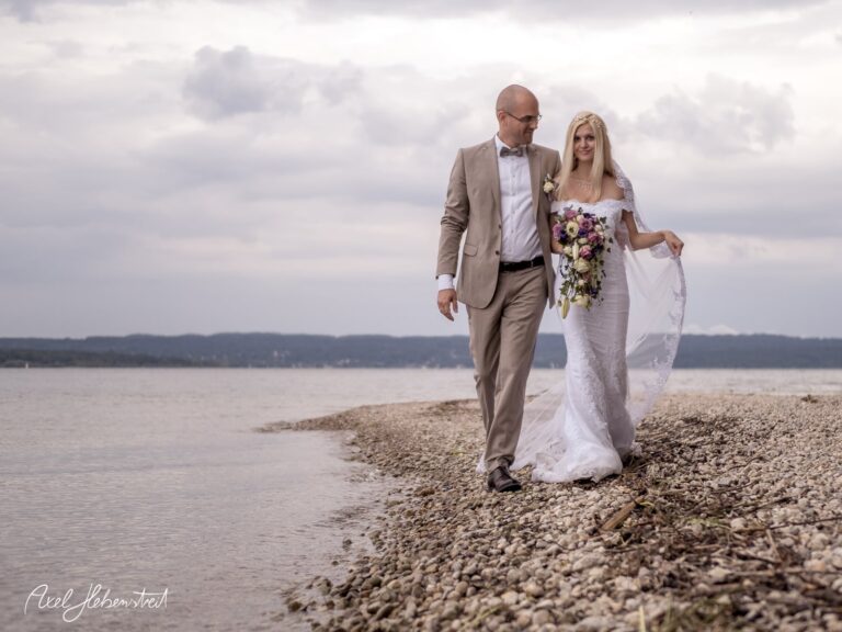 Brautpaar in eleganter Hochzeitskleidung spazieren am Ufer eines Sees und genießt den romantischen Moment – © Axel Hebenstreit
