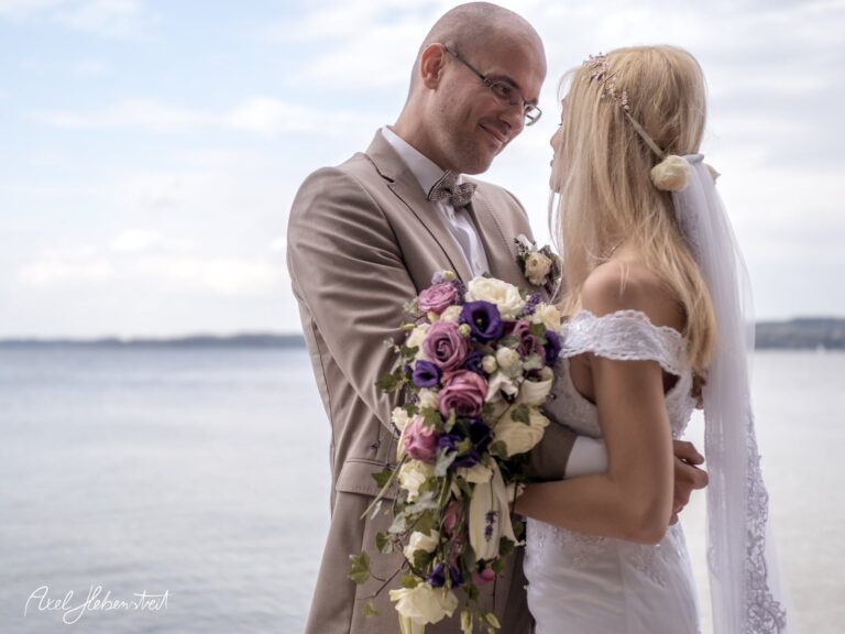 Brautpaar umarmt sich liebevoll am Wasser, umgeben von Blumen, in romantischer Hochzeitsatmosphäre – © Axel Hebenstreit