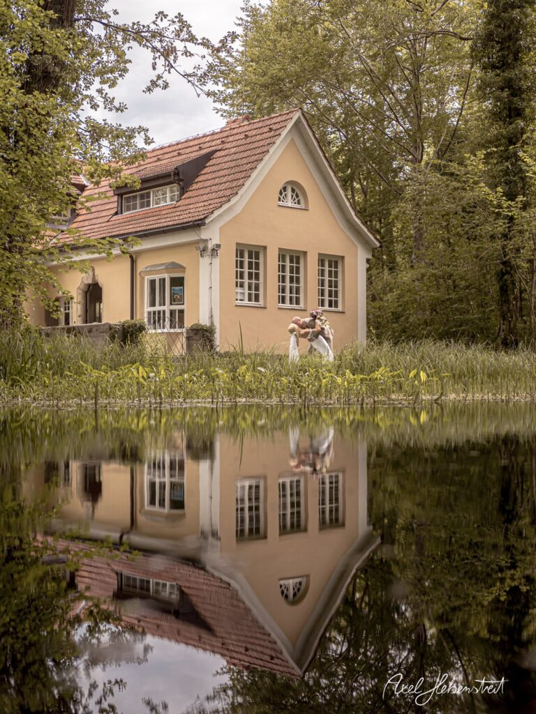 Ein Paar posiert romantisch vor einem charmanten Haus, dessen Reflexion im Wasser sichtbar ist – © Axel Hebenstreit