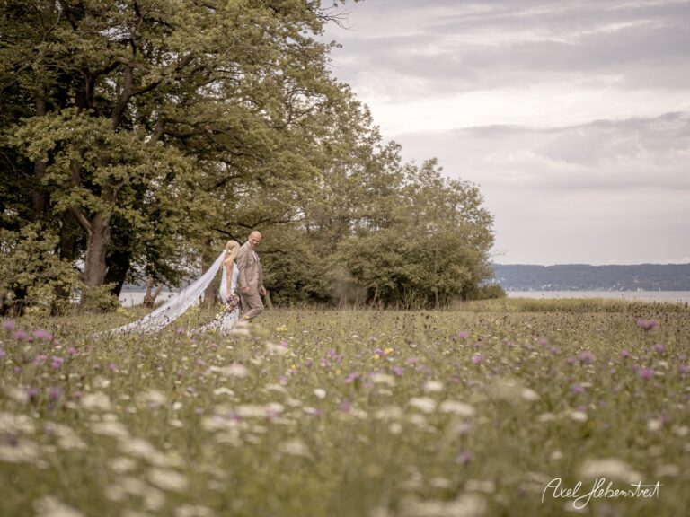 Hochzeitspaar in romantischer Pose auf blühender Wiese am See, umgeben von Natur – © Axel Hebenstreit