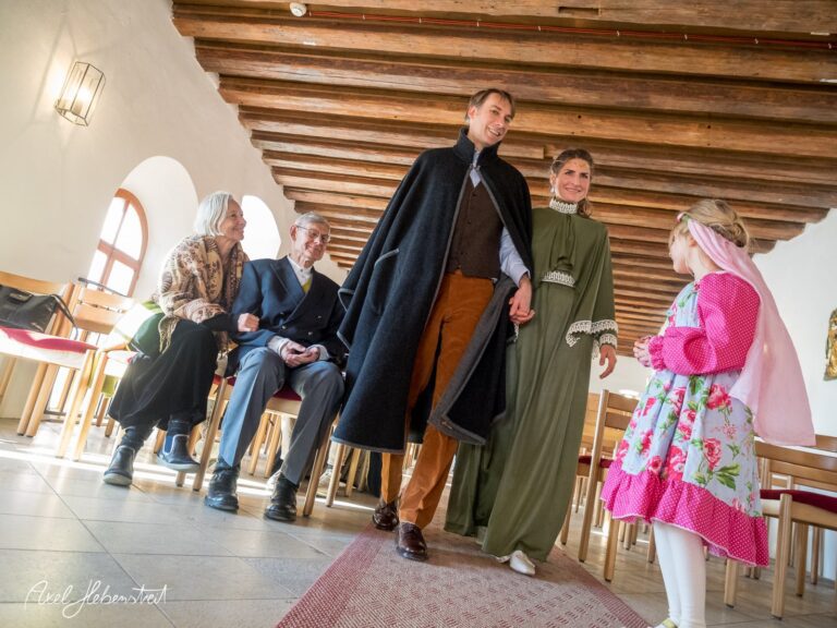 Eine Braut und ein Bräutigam in traditioneller Tracht schreiten in die Kirche, umgeben von Gästen in festlicher Stimmung. – © Axel Hebenstreit