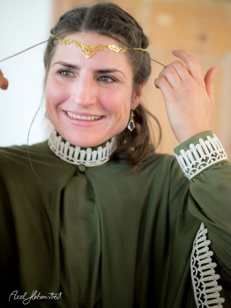 Eine lächelnde Frau in einem eleganten, grünen Gewand setzt ein goldenes Diadem auf – © Axel Hebenstreit.