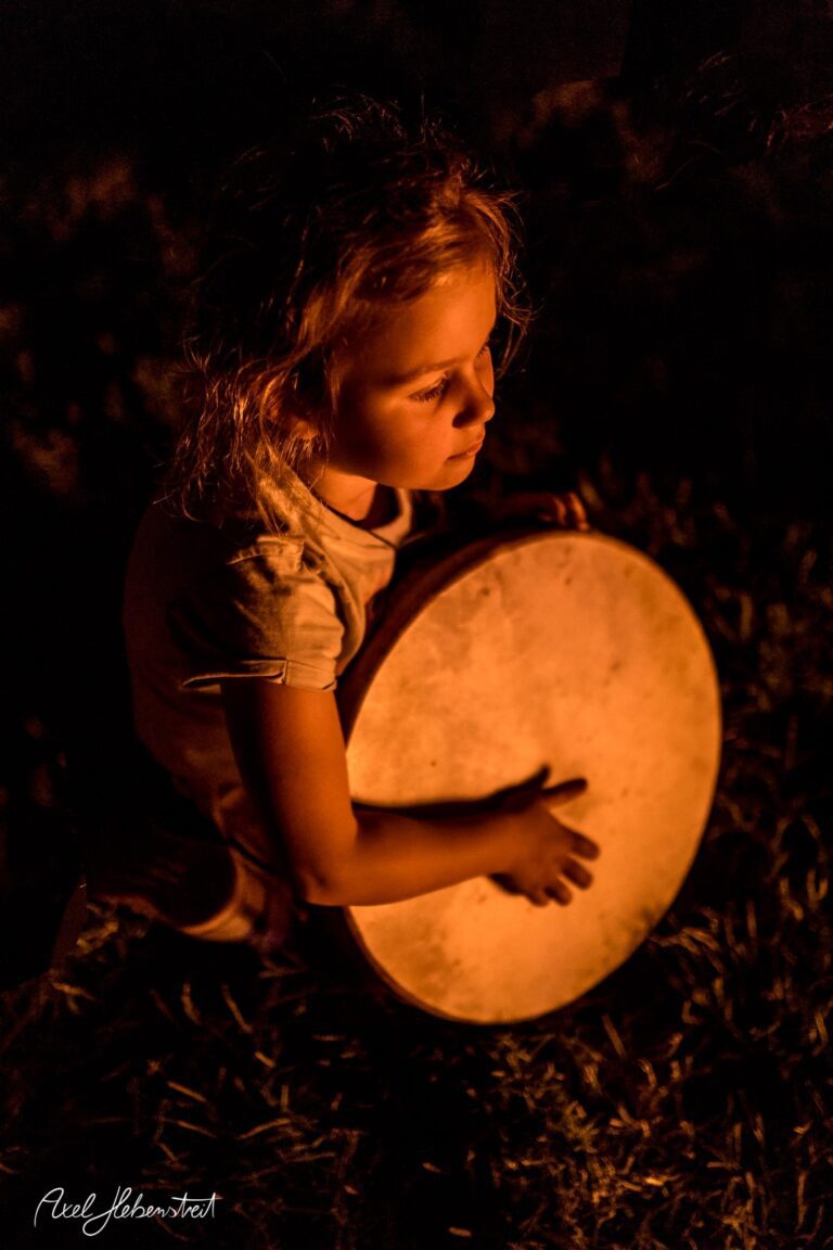Ein Mädchen spielt konzentriert auf einer Trommel, umgeben von warmem Licht und natürlicher Umgebung – © Axel Hebenstreit