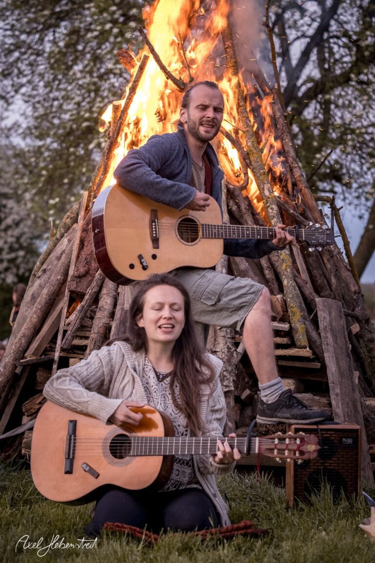 Musikende Menschen spielen Gitarre vor einem großen Lagerfeuer, umgeben von Natur – © Axel Hebenstreit