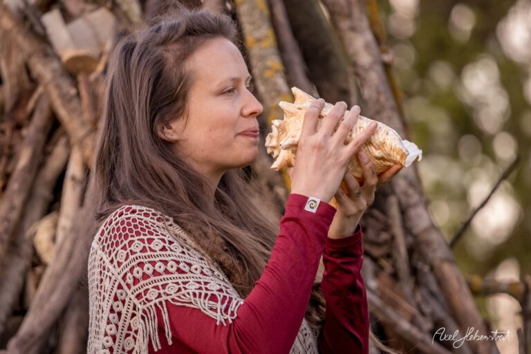Frau mit Muschel spielt ein melodisches Stück, umgeben von Natur und einem rustikalen Holzaufbau. – © Axel Hebenstreit
