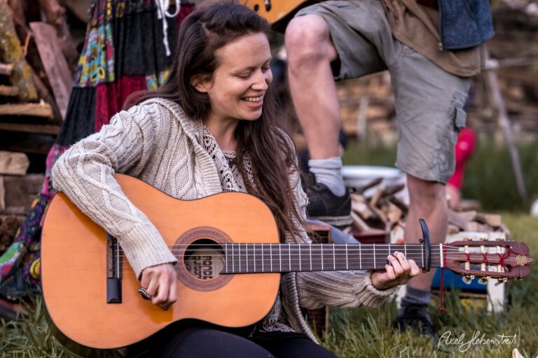 Eine lächelnde Frau spielt Gitarre in einer entspannten Festivalatmosphäre im Freien – © Axel Hebenstreit