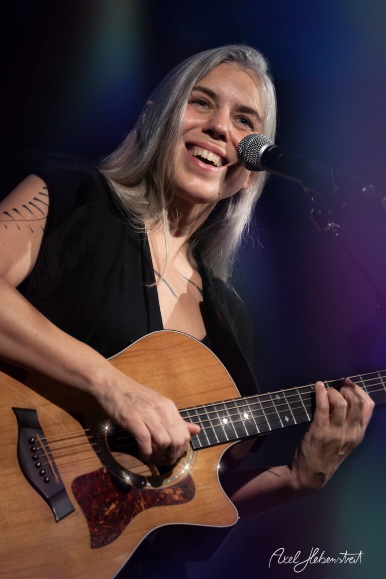 Eine lächelnde Sängerin mit Gitarre auf der Bühne, die ein einnehmendes Konzert gibt – © Axel Hebenstreit