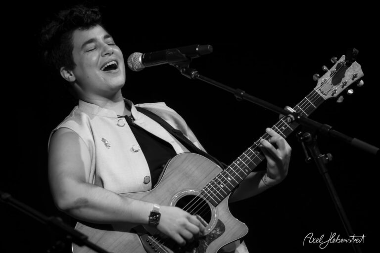 Eine Singer-Songwriterin, die mit Leidenschaft Gitarre spielt und dabei singt, vor einem dunklen Bühnenhintergrund – © Axel Hebenstreit