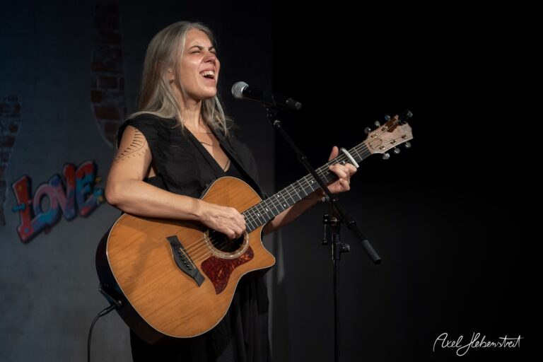 Eine Sängerin mit Gitarre performt leidenschaftlich auf einer Bühne, umgeben von einer lebendigen Kulisse. – © Axel Hebenstreit
