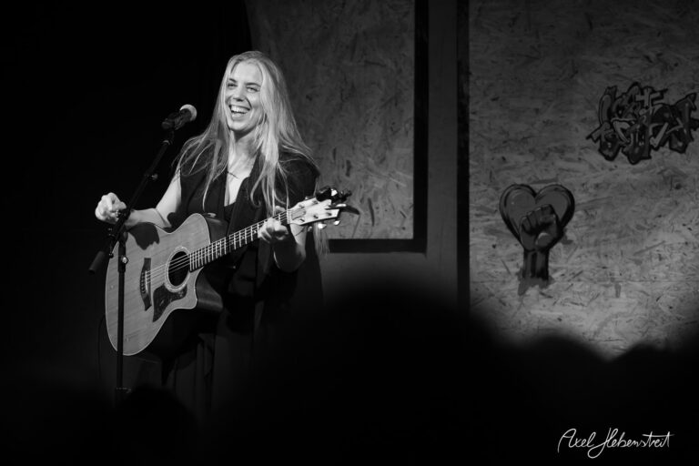 Eine Sängerin mit Gitarre lächelt fröhlich auf der Bühne und begeistert das Publikum – © Axel Hebenstreit.