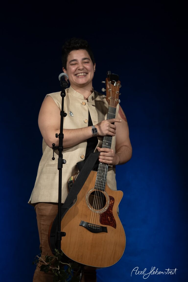Lächelnder Musiker mit Gitarre auf der Bühne, umgeben von einem stimmungsvollen blauen Hintergrund – © Axel Hebenstreit