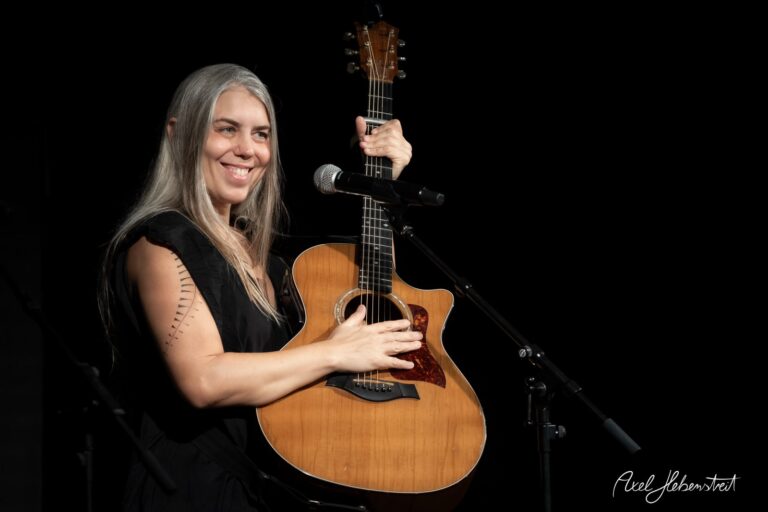 Eine lächelnde Musikerin mit Gitarre auf der Bühne, bereit für ihren Auftritt – © Axel Hebenstreit
