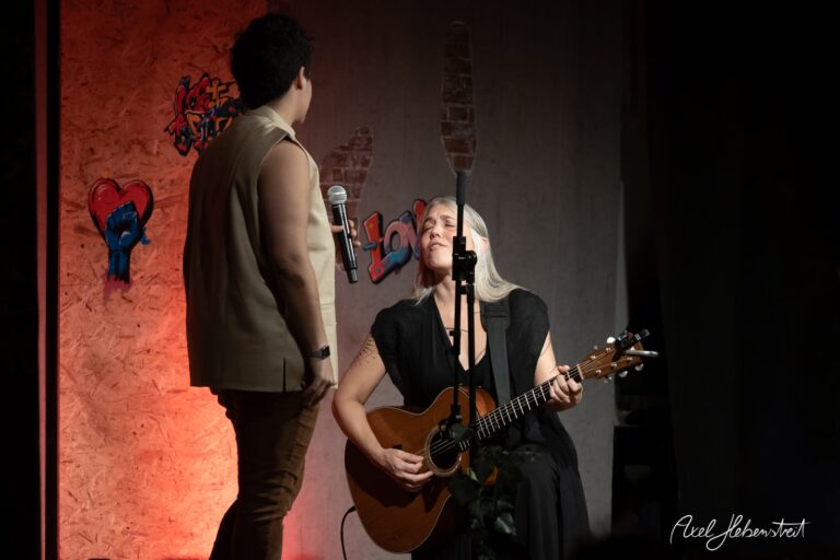 Zwei Musiker auf der Bühne, eine singt und spielt Gitarre, während der andere aufmerksam zuhört – © Axel Hebenstreit.