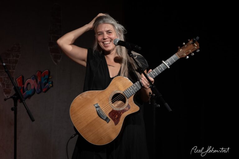 Frau mit Gitarre lächelt fröhlich auf der Bühne, umgeben von Mikrofonen und einer liebevollen Atmosphäre – © Axel Hebenstreit